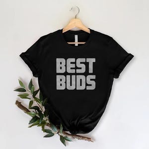 May include: Black t-shirt with the words "BEST BUDS" in a bold, textured, gray font. The shirt is hanging on a wooden hanger against a white background. A sprig of greenery is placed on the lower left side.