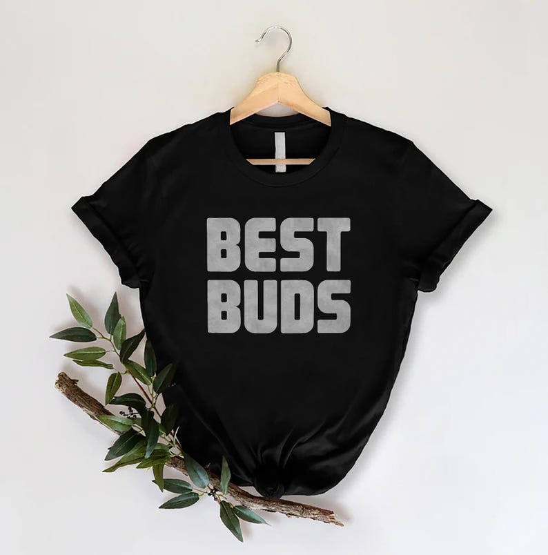 May include: Black t-shirt with the words "BEST BUDS" in a bold, textured, gray font. The shirt is hanging on a wooden hanger against a white background. A sprig of greenery is placed on the lower left side.