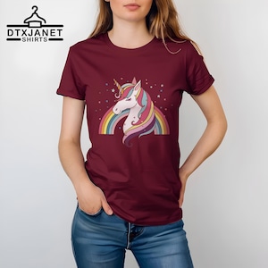 K&ouml;nnte beinhalten: Burgunderfarbenes T-Shirt mit einem bunten Einhorn-Design, Regenbogen und Sternen. Das Einhorn hat ein goldenes Horn und eine flie&szlig;ende M&auml;hne. Der Text "DTXJANET SHIRTS" befindet sich oben links.