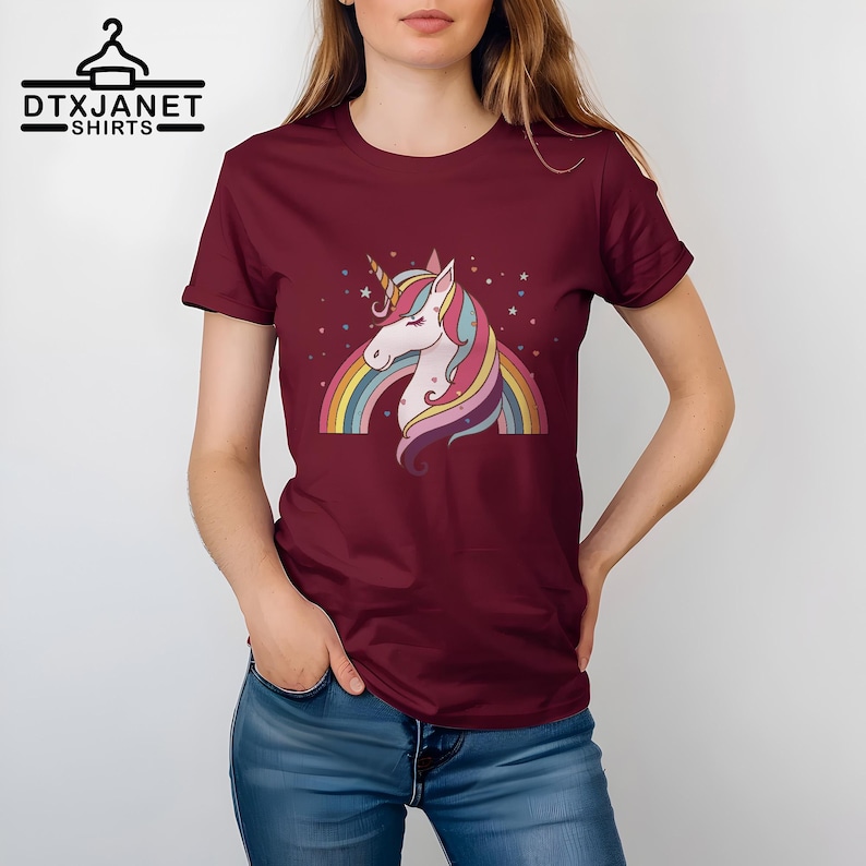K&ouml;nnte beinhalten: Burgunderfarbenes T-Shirt mit einem bunten Einhorn-Design, Regenbogen und Sternen. Das Einhorn hat ein goldenes Horn und eine flie&szlig;ende M&auml;hne. Der Text "DTXJANET SHIRTS" befindet sich oben links.