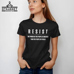 May include: Black t-shirt with the word "RESIST" in white block letters. Below the word "RESIST" is the text "THE POWER OF THE PEOPLE IS GREATER THAN THE PEOPLE IN POWER". The shirt has short sleeves.