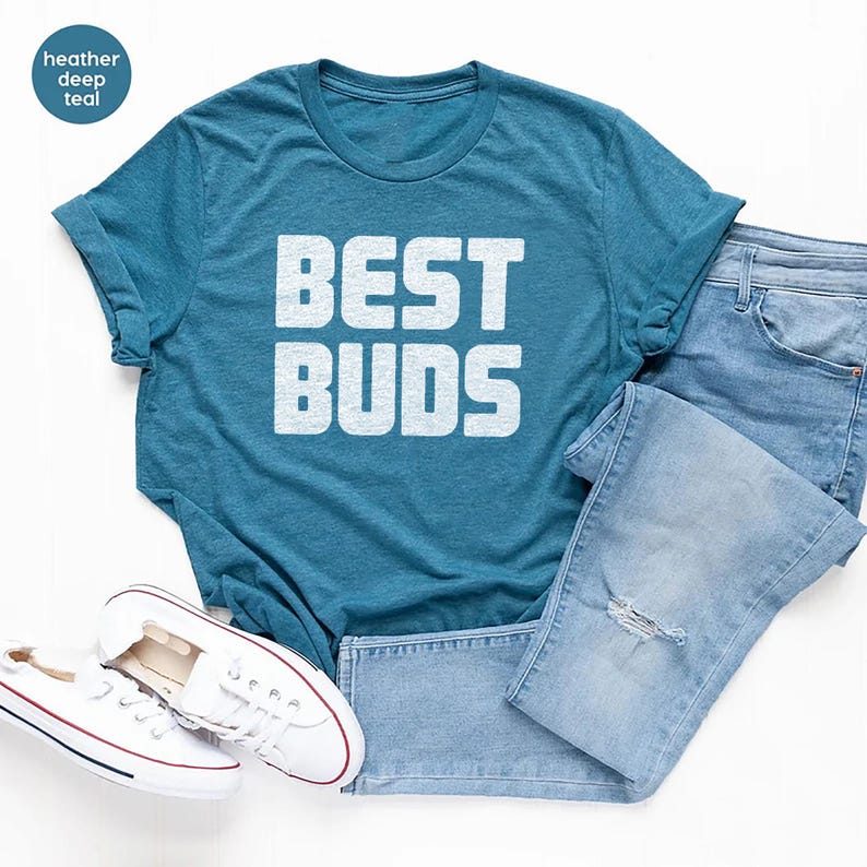 May include: A teal t-shirt with the text "BEST BUDS" in white block letters. The shirt is paired with light blue denim jeans and white sneakers with red accents. The image is a flat lay on a white surface.