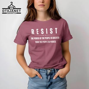 May include: A mauve t-shirt with the word "RESIST" in bold white letters. Below the word "RESIST" is the text "THE POWER OF THE PEOPLE IS GREATER THAN THE PEOPLE IN POWER". The shirt is a crew neck style.