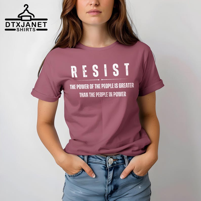 May include: A mauve t-shirt with the word "RESIST" in bold white letters. Below the word "RESIST" is the text "THE POWER OF THE PEOPLE IS GREATER THAN THE PEOPLE IN POWER". The shirt is a crew neck style.