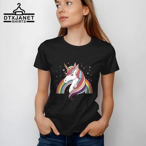 K&ouml;nnte beinhalten: Schwarzes T-Shirt mit einem bunten Einhorn-Design, Regenbogen und Sternen. Das Einhorn hat ein wei&szlig;es Gesicht, ein goldenes Horn und eine M&auml;hne in Rosa, Blau und Gelb. Der Text "DTXJANET SHIRTS" befindet sich oben links.
