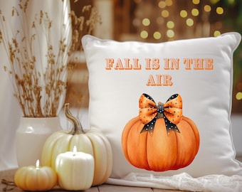 May include: White decorative pillow with the text "FALL IS IN THE AIR" and a watercolor pumpkin design. The pumpkin has a black bow with orange polka dots. A small pumpkin, candles, and a vase with dried plants are also visible.