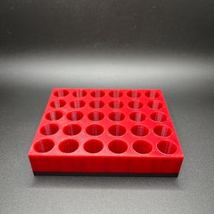 Puede incluir: Una bandeja cuadrada roja impresa en 3D con 42 agujeros circulares. La bandeja tiene una base negra y está diseñada para contener objetos cilíndricos pequeños. El fondo es negro liso.