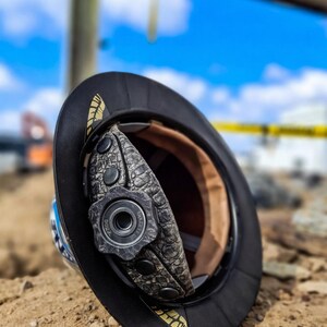 May include: A black hard hat with a textured gray interior suspension system featuring a central adjustment knob. The brim displays gold-colored feather-like accents. The interior lining is tan, and the adjustment dial is marked with the text Dura.