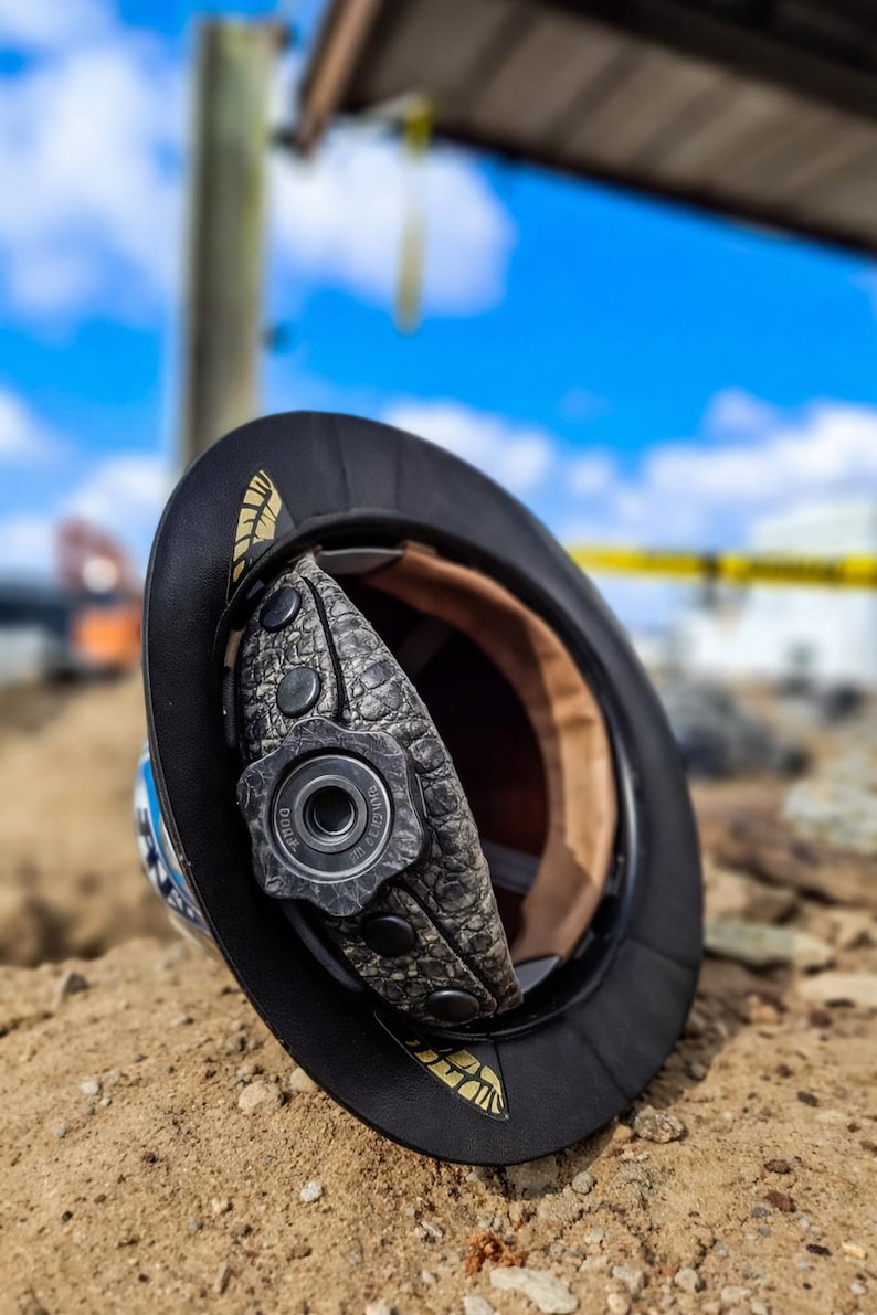 May include: A black hard hat with a textured gray interior suspension system featuring a central adjustment knob. The brim displays gold-colored feather-like accents. The interior lining is tan, and the adjustment dial is marked with the text Dura.