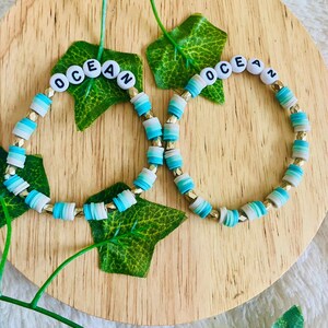 May include: Two beaded bracelets with the word "OCEAN" spelled out in white letters on blue and white beads. The bracelets are on a wooden surface with green leaves.