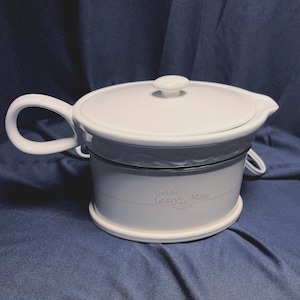 May include: A white ceramic Crock-Pot Gravy Mate warmer with a matching lid, a side handle, and a pouring spout. The base features the text Crock-Pot Gravy Mate printed in a light gray font. The appliance includes an attached electrical power cord.