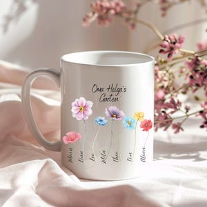 Peut inclure: Une tasse en céramique blanche avec un motif floral et le texte "Oma Helga's Garten" et les noms "Philine, Lisa, Lea, Lilla, Thea, Tina, Alissa" écrits sous les fleurs.