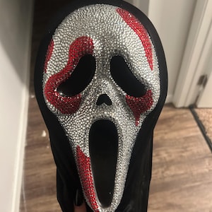 Op de afbeelding: Een Scream-masker met een zwarte capuchon. Het masker is bedekt met zilveren strass-steentjes, met rode strass-steentjes die de ogen en mond omlijnen. Het masker wordt door een persoon vastgehouden.