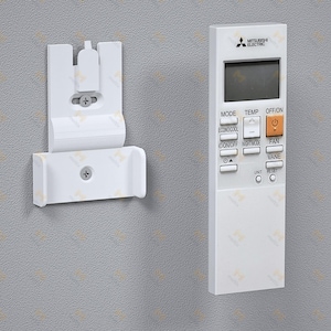 Op de afbeelding: Een witte Mitsubishi Electric airconditioner afstandsbediening en wandhouder. De afstandsbediening heeft een LCD-scherm en knoppen voor modus, temperatuur en ventilatorregeling. De wandhouder is ook wit.