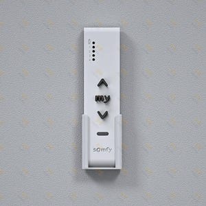 Op de afbeelding: Een witte Somfy afstandsbediening voor raambekleding, met een verticale rechthoekige vorm en een wandhouder. Het apparaat bevat zwarte knoppen voor omhoog, omlaag en een centrale 'my'-knop, samen met vier indicatielampjes gelabeld van 1 tot 4.