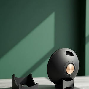 Puede incluir: Un altavoz esférico negro con un centro de color cobre y el texto "Harman Kardon". El altavoz está sobre un soporte negro, con un soporte negro separado a la izquierda. El fondo es verde oscuro liso.