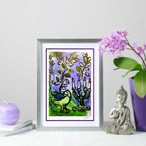 Op de afbeelding: Een ingelijste aquarel met een zilveren lijst, met een vogel, een kandelaar en bladeren in groen, blauw en paars. Een paarse kaars, een notitieboek en een kleine Boeddha-beeldje zijn ook aanwezig.
