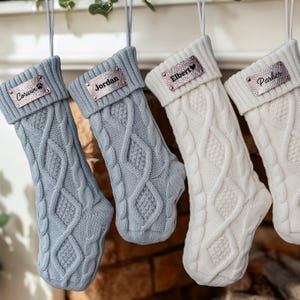 Peut inclure: Quatre bas de Noël en tricot torsadé de différentes nuances de gris et de blanc sont suspendus à une tringle décorative. Chaque bas a une étiquette nominative métallique personnalisée. Les noms sont "Corwin", "Jordan", "Elbert" et "Parker".
