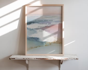 May include: A framed art print featuring a watercolor seascape in shades of blue, pink, and gold. The print includes the quote "I need the sea because it teaches me." by Pablo Neruda. The frame is light wood.