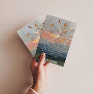 May include: Two art prints depicting geese in flight against a sunset. The prints include a quote about wild geese and the family of things. The prints are held by a person.