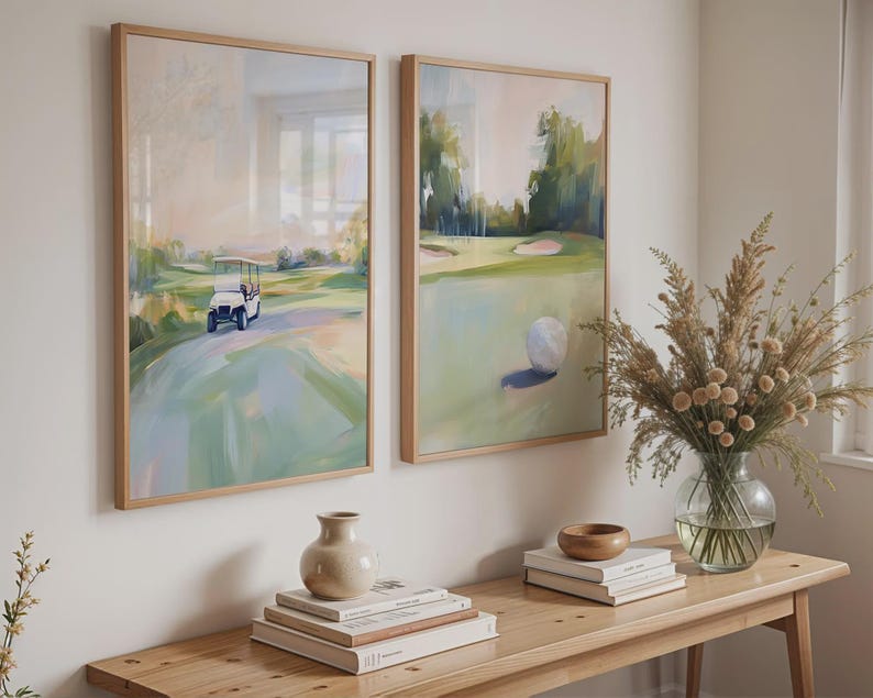 May include: Two framed abstract paintings depicting a golf course. The painting on the left shows a golf buggy driving on the course. The painting on the right shows a golf ball on the green. The paintings are hanging on a white wall above a wooden table with books and a vase of dried flowers.