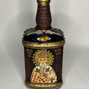 May include: A brown glass bottle with a gold top and a rope wrapped around the neck. The bottle is decorated with a gold trim and a painted icon of a saint. The icon has the text "Св. Сщмч. Феогн. Шк." in Cyrillic.