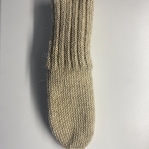 May include: A single, beige-colored knitted sock. The sock features a ribbed cuff and a textured, ribbed pattern throughout the leg and foot. The sock is made of a soft, natural fiber.