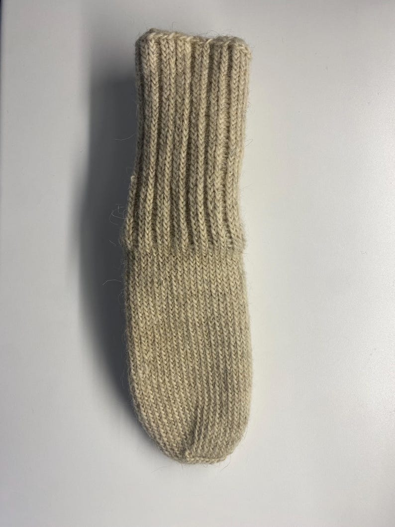 May include: A single, beige-colored knitted sock. The sock features a ribbed cuff and a textured, ribbed pattern throughout the leg and foot. The sock is made of a soft, natural fiber.