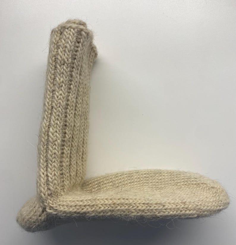 May include: A beige, hand-knitted sock with a ribbed leg and a rounded toe. The sock is made of a soft, textured yarn, and the stitches are clearly visible. The sock is lying flat on a white surface.
