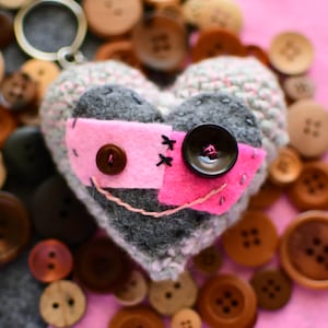 May include: A handmade heart-shaped keychain with a whimsical design. The heart is gray and pink, adorned with buttons and stitched details, resembling a face. It is surrounded by various wooden buttons on a pink background.