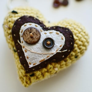 May include: Handmade yellow crocheted heart-shaped keychain with a brown and white fabric heart design. The fabric heart is adorned with two brown buttons and stitched details. A charming, unique accessory.