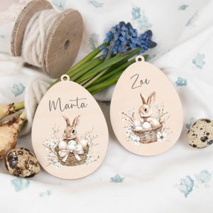 Op de afbeelding: Twee houten paasei-ornamenten met de namen "Marta" en "Zoe" erop gedrukt. Elk ei heeft een illustratie van een konijn in een mand met eieren en bloemaccenten. Een spoel wit garen en blauwe bloemen zijn op de achtergrond.