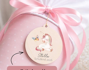 Pode incluir: Uma sacola de presente rosa com bolinhas brancas, amarrada com uma fita rosa, com uma etiqueta de madeira. A etiqueta apresenta uma ilustração de um unicórnio e uma borboleta, com o nome "Stella" e "Schulkind 2026".