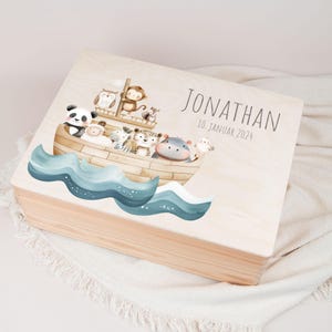 Könnte beinhalten: Eine Holzkiste mit einer hellen Holzoptik. Der Deckel zeigt eine Aquarell-Illustration eines Bootes mit Tieren darauf. Der Text "JONATHAN 10. JANUAR 2024" ist auf dem Deckel aufgedruckt.