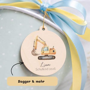Könnte beinhalten: Ein runder Holzanhänger mit einer Aquarellillustration eines gelben Baggers. Der Anhänger trägt den Namen "Liam" und den Text "Schulkind 2026". Er hängt an einem goldenen Faden, mit blauen und gelben Bändern im Hintergrund.