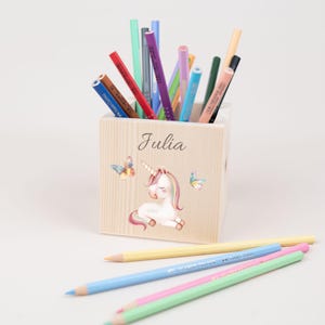 Puede incluir: Porta lápices de madera con un diseño de unicornio y mariposa. El porta lápices está personalizado con el nombre "Julia".