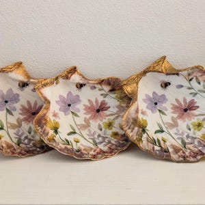 May include: Three decorative oyster shells with gold-painted edges, each featuring a hand-painted floral design with purple, pink, and yellow flowers, green leaves, and small bumblebees on a white interior surface, arranged in a horizontal row.