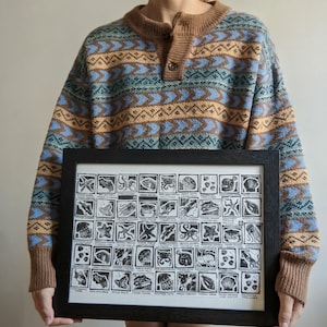 May include: A framed black and white linocut print featuring a grid of various marine life illustrations, each labeled with text. The artwork is displayed in a simple black frame, showcasing detailed carvings of sea creatures and shells.