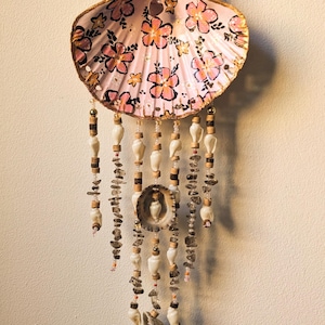 May include: A decorative wall hanging featuring a large, light pink scallop shell painted with black and orange floral patterns. Multiple strands of small white shells, brown wooden beads, and dark grey stone chips hang from the shell base.