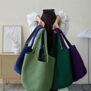 Op de afbeelding: Een collectie van vier handgemaakte gehaakte tote bags in effen kleuren, waaronder lichtgroen, donkergroen, diepblauw en paars. Elke tas heeft een gestructureerd geweven patroon, een open bovenkant en twee stevige schouderbanden om te dragen.