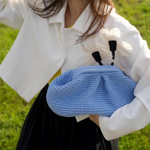 May include: A light blue crocheted clutch bag with a gathered top. The bag is held against a white jacket with flower accents. The jacket is worn over a yellow top and black skirt. The background is green grass.