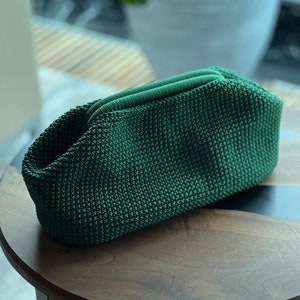 May include: A green knitted clutch bag with a textured surface. The bag is sitting on a wooden table with a black edge.