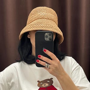 May include: A woman wearing a brown straw sun hat with a wide brim. She is wearing a white t-shirt with a cartoon bear wearing a red jacket and holding an umbrella. The text on the shirt reads "Beauty is superhic,  aeeop yly vtyuty".