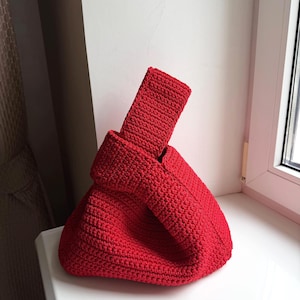 Op de afbeelding: Een felrode gehaakte handtas met een gestructureerd knoophandvat. De tas is gemaakt met een dicht, getextureerd steekpatroon, wat zorgt voor een solide en uniform uiterlijk over het gehele buitenoppervlak van dit kleine accessoire.