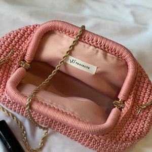 May include: A pink crocheted clutch bag with a gold chain strap. The bag has a clasp closure and a small label inside that reads "W PROJECTS".