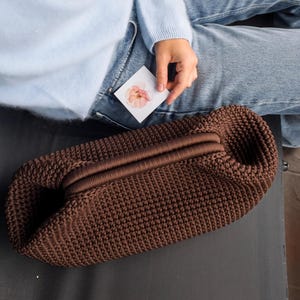 May include: A brown, hand-crocheted clutch bag with a rounded shape. The bag features a textured stitch pattern and a central, horizontal band. The bag is resting on a dark surface.