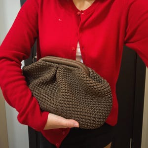 May include: A red cardigan with a brown crocheted clutch. The clutch has a textured, woven appearance and a brown handle. The cardigan is buttoned and has a classic, simple design. The overall style is casual and chic.
