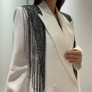 May include: A white blazer with a double-breasted design and a wide lapel. The blazer features black fringe and sequin detailing across the shoulders and down the front, adding a decorative element to the garment.