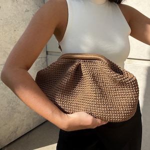 May include: A brown, textured clutch bag with a woven design. The bag has a rounded shape and a contrasting brown top. The bag is held against a white top and black pants.