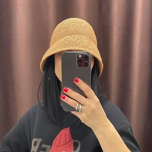 May include: A light brown woven straw bucket hat. The hat is worn by a person wearing a black t-shirt with a graphic design of a bear wearing a red beanie. The person is holding a black smartphone and has red nail polish.